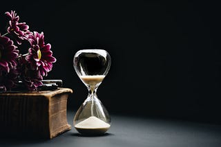 Photo of an hourglass with sand running out, juxtaposed to a thick book with purple flowers on top.