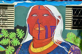 A colourful wall mural of an elder’s face. They have long white hair and markings on their left cheek and from nose to chin in purple.