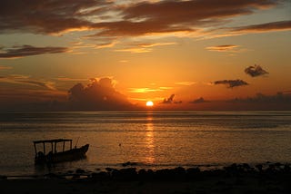 The sun peeking up on the horizon, over an ocean with a humble boat anchored at the rocky shore.