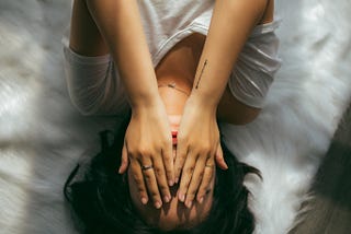 An aerial view of a woman in a white tee shirt laying on a crumpled white sheeted bed, her hands covering her face.