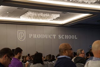 Conference room filled with people sitting down for a session with the Product School visible on a grey wall in the distance.