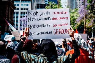 Protester holding sign that reads, “Not war, its colonialism. Not eviction, its ethnic clensing. Not conflict, it’s occupation. Not complicated, it’s GENOCIDE”.
