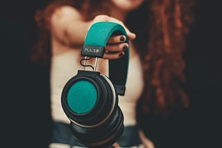 a woman with her arm extended toward the camera holding a pair of headphones as if offering them to you