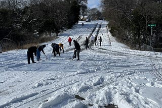 15 people wrapped in snow gear with shovels in hand shovveling 8 inches of snow from steep roads.