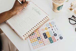 A planner and colorful stickers on a desk, with a human hand holding a pencil, about to write. A Starbucks iced coffee and a pair of eyeglasses are also on the desk.
