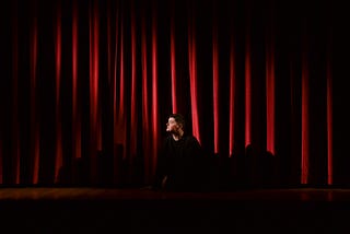 A person sitting behind the closed red curtain on a stage, looking to his right.