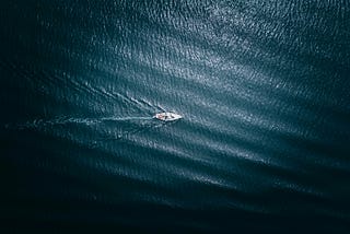 A small sailboat moves alone through dark blue ocean waters, leaving ripples behind it.