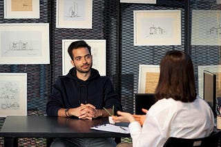 brown man being interviewed across a table in a room of architectural drawings