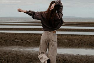 A woman stands on one foot, one arm extended, the other crooked behind her head. She faces away from us, towards streaks of land and water. She wears a dark sweater and beige long pants.