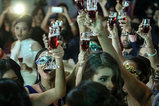 Party scene with a group of people raising glasses filled with red alcohol in the air. In the background, guests are filming on their phones and in the foreground, you can see part of a lady’s face, but she looks distressed.