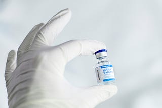 A close-up of a person’s hand wearing a white latex glove, gently holding a small glass vial between thumb and forefinger against a soft, light blue-gray background. The vial has a blue-and-silver cap and a white label reading “SARS-CoV-2 mRNA Vaccine COVID-19,” with “EXP 2021–06–03” printed vertically along the side.