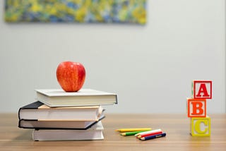 Red apple sitting on a stack of four books on a wooden tabletop, next to colored pencils and three wooden ABC blocks. The background is blurred.