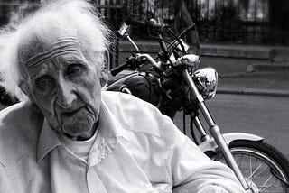 An elderly man sits in front of a motorcycle with his eyes downcast looking regretful