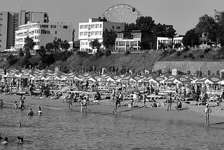 A black and white view of a beach, taken from the sea. The sand is crowded with sunshades; low-rise apartment blocks or hotels stand in the background.