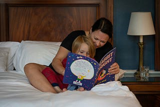 woman with arms around child on bed reading a book to him