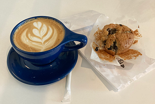 a latte in a blue mug next to a partially eaten muffin