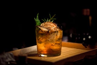 An alcoholic drink with herbs sticking out of it on a wooden tray.