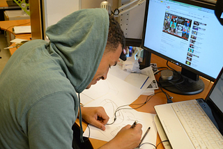 A Black male designer in a hooded sweatshirt sketches on paper while in front of his computer.