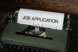 A typewriter with a piece of white paper with the words JOB APPLICATION in bold, all caps font typed on it.