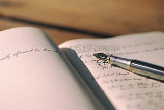 An ink pen laying on top of an open notebook. The notebook has writing in it.