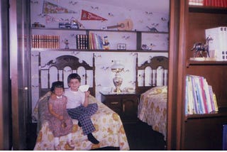 A little girl of 3 is sitting on the edge of a bed. Her older brother is hugging her and holding a paper airplane.