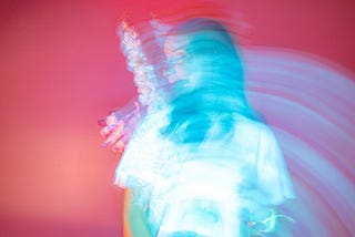 A woman poses with an orchid; she is artistically blurred by multi-exposure phtotography. The photo features a rich palate of red, orange, violet, and cool blues.