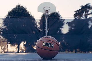 A basketball in the foreground where NBA next to the NBA logo is clearly seen. In the background is a park or playground outdoor basketball hoop against a backdrop of trees.