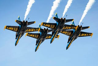 four U.S. Navy blue angels aircraft in flight in blue sky. 4 navy jet aircraft with yellow text that says U.S. Navy with white jet stream from aircraft; posted on Mickey Markoff 2024 article on U.S. Navy birthday