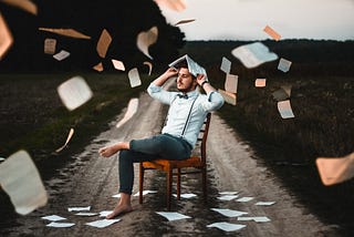 A barefoot man dressed in a nice shirt is sitting on a chair in the middle of dirt path. He has an open book over his head and there are loose pages fluttering around everywhere in the image.