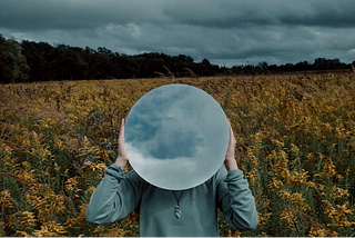 A person in a blue top standing in a field full of yellow flowers. The sky is overcast and grey. In place of a head, the figure holds a circular mirror up that reflects the sky above.