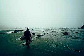 A person dressed in all dark clothing walks on a desolate beach alone.