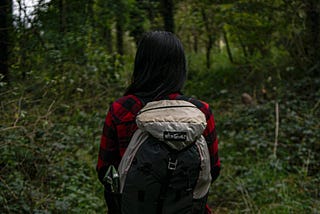 Person with long black hair standing in a forest. Wearing a hiking backpack, and red flannel shirt.