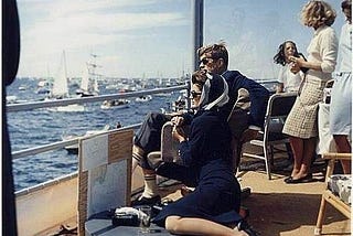 A picture of JFK and Jackie sitting in a ship looking out at sailboats and small water craft on a bright sunny day.