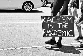 Person holding a sign that reads “Racism is the pandemic”.