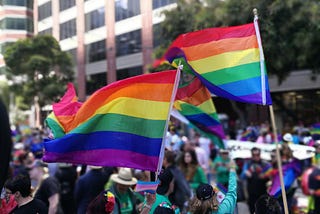 A crowd of people on a street, none clearly visible, with three rainbow flags in the foreground.