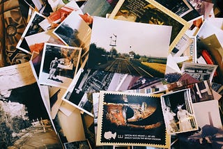 A scattered collection of old photographs and postcards in a mix of color and black-and-white, spread across a surface. The photos include scenes of people, landscapes, railways, and historical artifacts, with one prominent postcard showing the Sutton Hoo helmet from the British Museum. The arrangement evokes a nostalgic and archival feeling, capturing moments from different times and places.