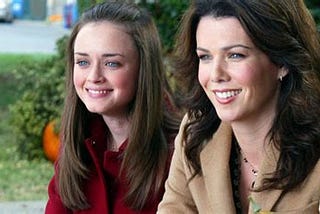 An image of the characters Rory Gilmore and her mother, Lorelai Gilmore, from the show “Gilmore Girls.” They are both white women with longish brown hair and blue eyes. Rory on the left is wearing a red coat and her hair pulled back from her forehead. Lorelai is wearing a camel coat and a necklace and has her hair swept back. They are smiling at something to the left off-camera.