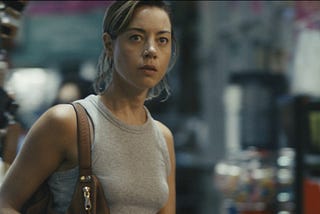A white woman in her thirties stands in a warehouse with a blurred background. Her brown hair is pulled back. She wears a gray tank top and has a brown shoulder bag.