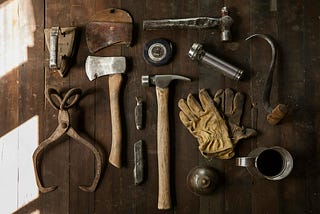 A variety of tools, including an axe, two hammers, a flashlight, and a pair of gloves, among others, laid out in a neat fashion.