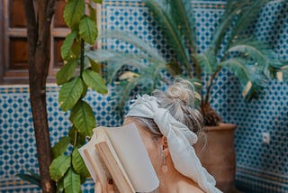 A woman sitting with her face covered behind a book, and a mobile in her hand