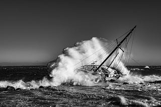 Black and white photo of an abandoned sailboat with a wave crashing over it.