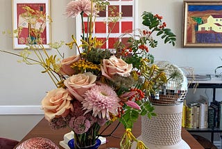 A large sunny bouquet of flowers, including pink roses and other pink and red flowers. The bouquet is on a table surrounded by small disco balls, and paintings are on the wall behind them.