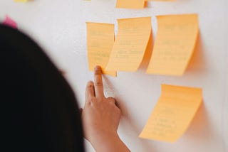 Uma mulher apontando para um quadro de post-its de cores laranja e rosa.