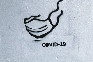 Graffiti on a white wall displaying a mask and “COVID-19” written below it.