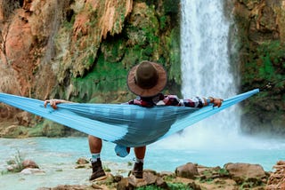 A man sitting in a hammock facing away from the camera and towards a waterfall.