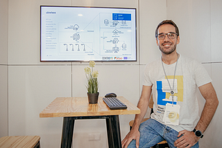 A man sitting on a stool in front of a presentation screen at a Smart City Expo, smiling and wearing a conference badge, with a computer keyboard on the table in front of him. The screen behind him displays a diagram related to smart city technology.
