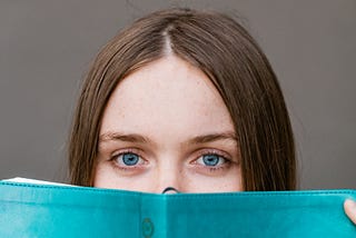 A white woman with brown hair and blue eyes peers out to look at the camera from behind a blue book that she is holding.