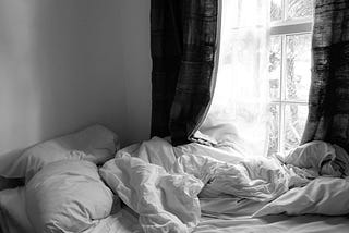 A black and white photo of an unmade bed. The bed has white covers, and is against a window with black curtains and white voile. The duvet is crumpled and the pillows strewn haphazardly on the left.