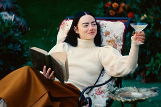 Emma Stone as Bella Baxter in Poor things, sitting on a chair and toasting a martini whilst reading a book