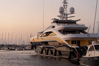 Photo of a large yacht at a dock with smaller boats nearby. The large yacht has three levels visible above the water line, with a deck on each level. The sun is setting in the background, with a hazy blend of white, yellow, pink and purple.